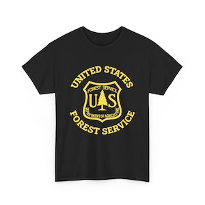May include: A black short-sleeved t-shirt featuring a yellow graphic print on the chest. The design displays the United States Forest Service shield emblem with a central pine tree icon, the letters U.S., and the text United States Forest Service.