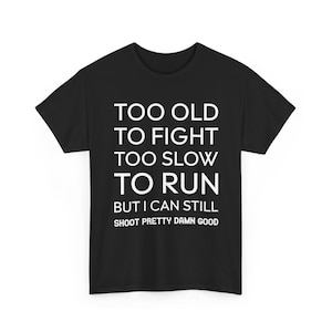 Op de afbeelding: Zwart T-shirt met witte tekst. De tekst luidt: "TOO OLD TO FIGHT TOO SLOW TO RUN BUT I CAN STILL SHOOT PRETTY DAMN GOOD."