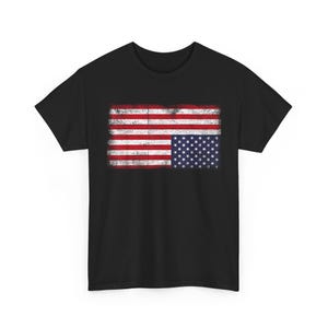 Puede incluir: Camiseta negra con un diseño desgastado de la bandera estadounidense. El gráfico de la bandera incluye rayas rojas y blancas y un campo azul con estrellas blancas. La camiseta es de cuello redondo.