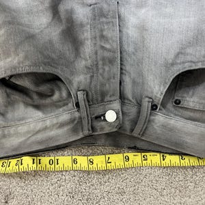 May include: Close-up of gray denim jeans with a button closure and pockets. A yellow measuring tape shows a length of approximately 15 inches. The jeans have a faded, worn appearance.