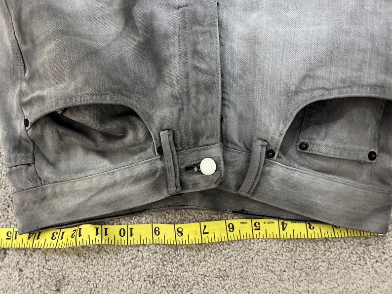 May include: Close-up of gray denim jeans with a button closure and pockets. A yellow measuring tape shows a length of approximately 15 inches. The jeans have a faded, worn appearance.