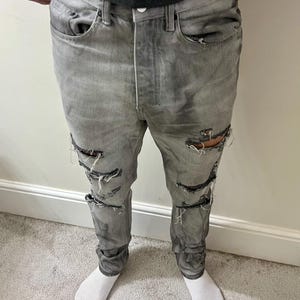 May include: Gray denim jeans with multiple distressed rips along the legs. The jeans have a button fly and classic five-pocket design. The fabric appears worn and faded, giving them a vintage look. The person is wearing white socks.