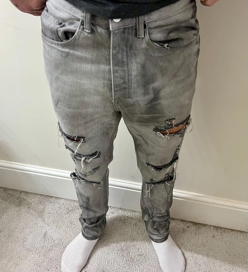 May include: Gray denim jeans with multiple distressed rips along the legs. The jeans have a button fly and classic five-pocket design. The fabric appears worn and faded, giving them a vintage look. The person is wearing white socks.