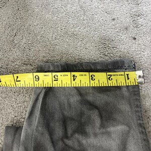 May include: Close-up of gray denim jeans with a yellow measuring tape showing a measurement of approximately 6 inches. The jeans are folded, and the tape is placed across the fabric to measure the width.