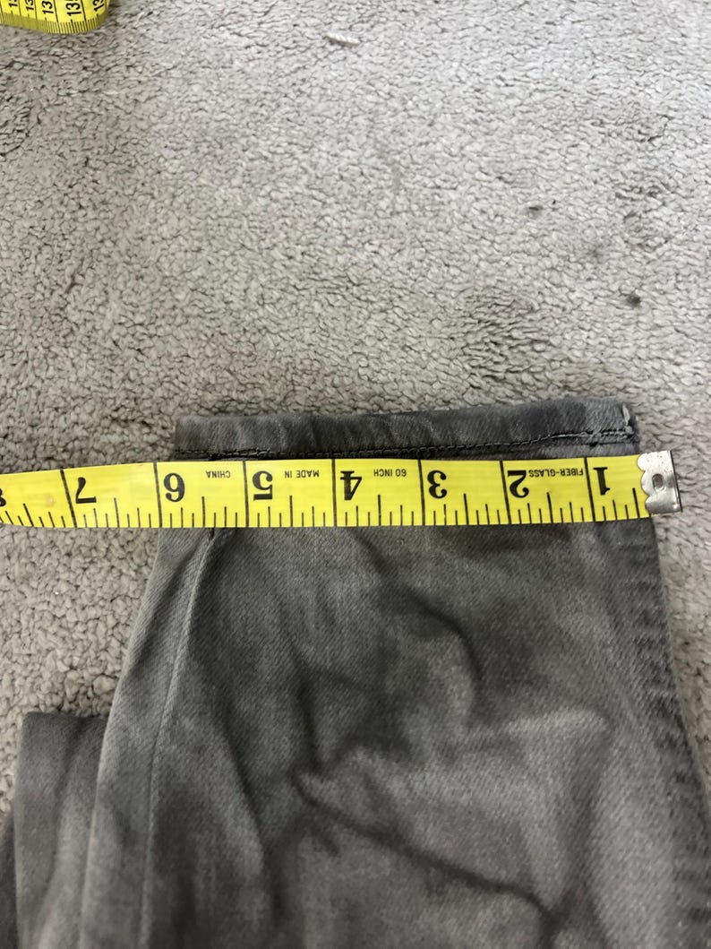 May include: Close-up of gray denim jeans with a yellow measuring tape showing a measurement of approximately 6 inches. The jeans are folded, and the tape is placed across the fabric to measure the width.