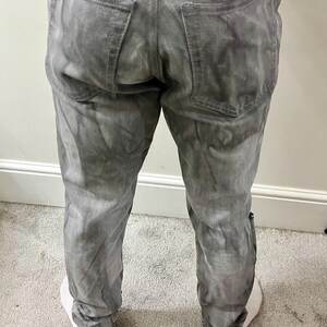 May include: Gray denim jeans with a faded, textured appearance. The jeans have a classic five-pocket design, with two back pockets and two front pockets, plus a small coin pocket. The jeans are worn with white sneakers.