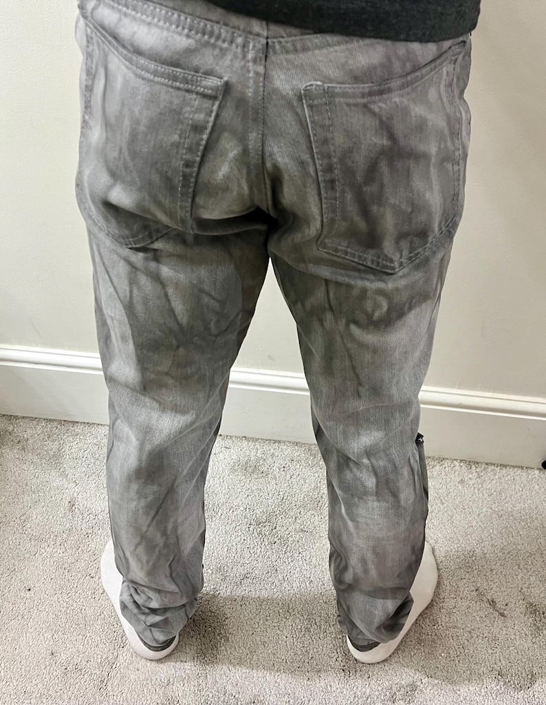 May include: Gray denim jeans with a faded, textured appearance. The jeans have a classic five-pocket design, with two back pockets and two front pockets, plus a small coin pocket. The jeans are worn with white sneakers.