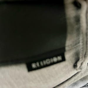May include: Close-up of a gray garment with the word "RELIGION" printed in white on a black rectangular label. The garment appears to be made of a soft, textured fabric.