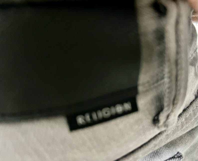 May include: Close-up of a gray garment with the word "RELIGION" printed in white on a black rectangular label. The garment appears to be made of a soft, textured fabric.
