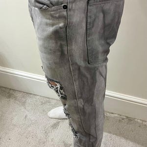 May include: Gray denim jeans with distressed details. The jeans feature ripped sections on the knees and thighs, with visible white fabric underneath. The jeans have pockets and a button closure.