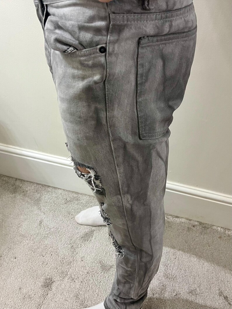 May include: Gray denim jeans with distressed details. The jeans feature ripped sections on the knees and thighs, with visible white fabric underneath. The jeans have pockets and a button closure.