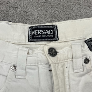Puede incluir: Primer plano de unos pantalones vaqueros blancos con la etiqueta "Versace Jeans Couture" visible. Los pantalones tienen cierre de botón y herrajes metálicos decorativos. La tela parece ser una mezcla de algodón resistente.