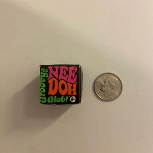 Può includere: Una piccola scatola nera del giocattolo antistress "The Nee Doh Groovy Glob", con testo in rosa, arancione e verde. Una moneta è accanto alla scatola per il confronto delle dimensioni. Il giocattolo è per bambini dai 3 anni in su.