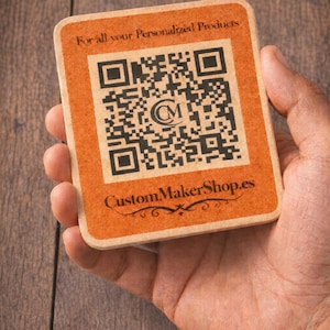 May include: A rectangular, orange-toned card with a QR code and the text "For all your Personalized Products" and "CustomMakerShop.es". The card is held in a hand, with a wooden background.
