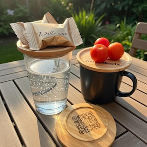 May include: A clear glass with a bamboo lid and a gold-wrapped candy with the word "Delizze" on top. A black mug with a bamboo lid and three red tomatoes. A bamboo coaster with a QR code.
