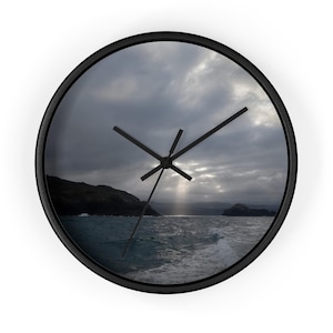 May include: A round wall clock with a black frame featuring a photographic print of a dark, cloudy seascape. The image displays a rocky coastline, turbulent ocean waves, and a single beam of light breaking through the grey sky. Three black hands.