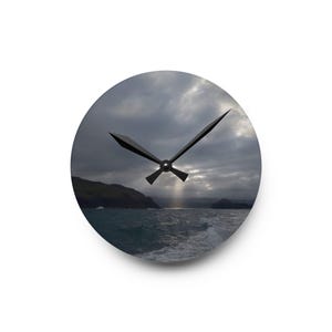 May include: A circular wall clock featuring a photographic print of a dark, cloudy seascape with rocky cliffs and a sunbeam breaking through the clouds. The clock has two black, pointed hands centered on the image of the ocean and sky.