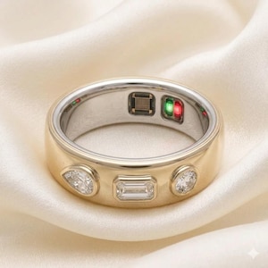 Può includere: Un anello smart dorato con tre gemme trasparenti incastonate a taglio pera, smeraldo e rotondo. L'interno della fascia mostra una serie di sensori metallici e un piccolo modulo rettangolare con spie luminose verdi e rosse accese.