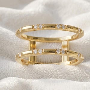 Peut inclure: Deux bagues dorées reliées par des barres verticales. Chaque bague est ornée de petites pierres scintillantes. Les bagues sont posées sur un fond blanc doux, créant une esthétique luxueuse et élégante.