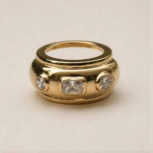 May include: A wide gold band ring featuring a polished finish with three embedded clear stones. The centre stone is rectangular, flanked by two smaller stones, one round and one pear-shaped, all set flush into the thick, rounded metal surface.
