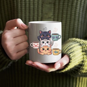 May include: White ceramic mug with a design of three cartoon cats, each holding a coffee cup. The cups feature heart, paw print, and fish motifs. Steam rises from the cups, suggesting hot drinks.