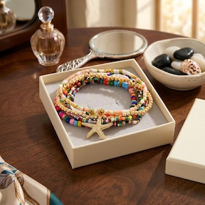 Puede incluir: Un conjunto de pulseras de cuentas coloridas con detalles dorados y un dije de estrella de mar, presentado en una caja color crema. Las pulseras presentan una variedad de colores, incluyendo rosa, azul, verde y amarillo. Sobre una superficie de madera oscura.