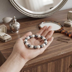 Puede incluir: Una pulsera circular compuesta por cuentas esféricas lisas que alternan entre blanco y gris oscuro. Las cuentas están ensartadas en un patrón repetitivo, creando una forma redondeada y uniforme que descansa en el centro de una palma abierta.