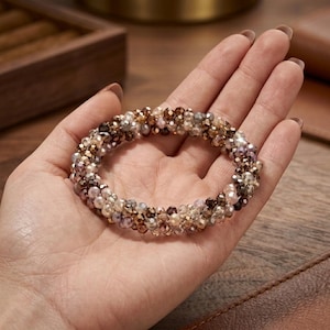 Op de afbeelding: Een armband van kleine, veelkleurige kralen in bruin-, goud- en paarstinten. De armband wordt op een hand getoond, waarbij het gedetailleerde ontwerp en de kleurvariaties worden benadrukt. De kralen zijn rond en creëren een textuur.