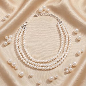 May include: A three-strand pearl necklace with a silver clasp, displayed on a cream-colored satin fabric. Scattered around the necklace are various sizes of individual pearls, creating an elegant and classic aesthetic.