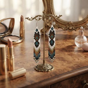 May include: A pair of long, dangling earrings crafted from small seed beads in a geometric pattern. The design features a palette of white, black, brown, and teal colors, with a fringe of loose beads at the bottom of each earring.