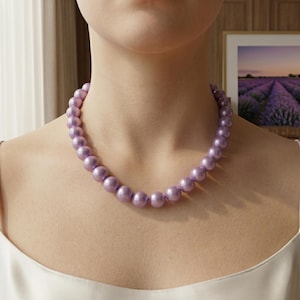 Könnte beinhalten: Eine lavendelfarbene Perlenkette. Die Halskette besteht aus großen, runden Perlen, die einen klassischen und eleganten Look erzeugen. Die Perlen sind gleichmäßig angeordnet und haben eine glatte, glänzende Oberfläche. Die Halskette wird um den Hals getragen.