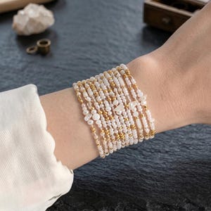 May include: A stack of beaded bracelets featuring white pearls and gold-colored beads. The bracelets are worn on a wrist, showcasing the delicate design and varying bead sizes. The bracelets have a mix of small and larger beads.