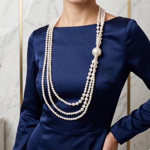 Puede incluir: Un collar de perlas de varias hebras con un gran acento de perla. El collar se usa sobre un vestido azul marino. El fondo es una pared de mármol con detalles dorados. Las perlas son blancas y el vestido es de color sólido.