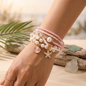 Pode incluir: Um conjunto de pulseiras de contas em tons de branco, rosa e dourado. As pulseiras apresentam contas de pérolas, pequenas contas de sementes e berloques, incluindo uma concha e uma estrela do mar. As pulseiras são usadas no pulso.