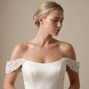 May include: A white off-the-shoulder gown featuring delicate, textured floral embellishments on the sleeves. The bodice is crafted from a smooth, solid fabric with a clean neckline, highlighting the intricate beadwork and floral detailing on the shoulders.