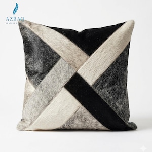 May include: A square throw pillow featuring a geometric patchwork design made of hair-on hide. The pattern consists of alternating diagonal strips in solid black, off-white, and mottled grey tones, creating a cross-like motif across the surface.
