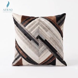 May include: A square throw pillow featuring a patchwork design of genuine cowhide leather. The surface displays a geometric chevron pattern composed of alternating strips in shades of white, cream, gray, dark brown, and black hair-on hide.