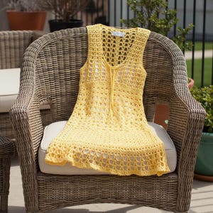 May include: A sleeveless, yellow crochet dress with a V-neck and a drawstring closure. The dress is made of a lightweight, open-weave yarn, and is displayed on a wicker chair with a white cushion. The dress has a scalloped hemline.