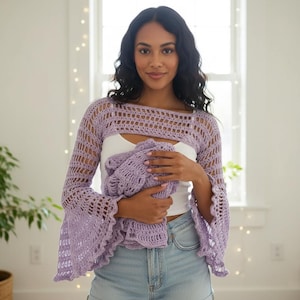May include: A light purple crochet shrug with bell sleeves. The open-weave design features a cropped top and long sleeves. The woman in the image is wearing the shrug over a white top and denim shorts. The shrug is made of a soft, lightweight yarn.