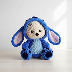 May include: A handmade plush toy in the shape of a bunny, featuring a blue hooded outfit and large ears with pink inner lining. The bunny has a cream-colored face with black eyes and a black nose. The toy is made of soft, textured yarn.