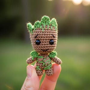 Puede incluir: Una figura de ganchillo de un personaje con cuerpo marrón, detalles verdes y ojos negros. La figura tiene una expresión sonriente y se sostiene en una mano. La cabeza y los hombros de la figura son verdes, y el cuerpo tiene detalles verdes en forma de hoja.