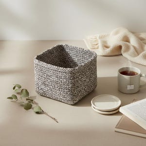 Könnte beinhalten: Ein quadratischer, grau-weißer Häkelkorb steht auf einer hellen Oberfläche. Eine Tasse Tee, zwei kleine weiße Untertassen, ein Buch und ein Zweig Grünzeug sind ebenfalls vorhanden. Eine cremefarbene Decke befindet sich im Hintergrund.