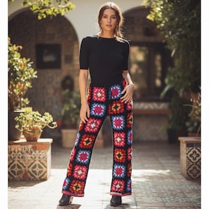 May include: A woman wearing a black short-sleeve top and colorful crochet pants. The pants feature a black background with square patterns in red, pink, blue, and yellow. The pants have a drawstring waist.