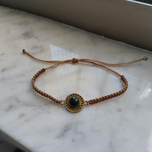 Può includere: Un bracciale in corda marrone con un ciondolo centrale circolare in ottone con un inserto in pietra verde scuro. Il bracciale è realizzato con un design a corda intrecciata e include cordini regolabili su ciascun lato per fissare la misura.