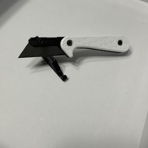 Könnte beinhalten: Ein Cuttermesser mit schwarzer Klinge und weißem Griff. Der Griff hat ein strukturiertes geometrisches Muster und ist mit zwei schwarzen Schrauben befestigt. Die Klinge ist ausgefahren und einsatzbereit.