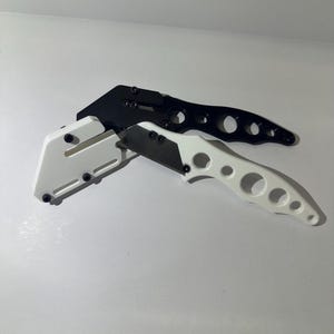 Puede incluir: Se muestran dos cuchillos utilitarios, uno negro y otro blanco. El cuchillo negro está detrás del blanco. Ambos cuchillos tienen un mango curvo con recortes circulares y una hoja recta. El cuchillo blanco tiene un mango blanco y una hoja negra.