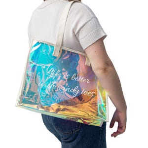 May include: Iridescent tote bag with white handles and trim. The bag features the text "Life is better with sandy toes" in white script. The bag is carried over the shoulder.