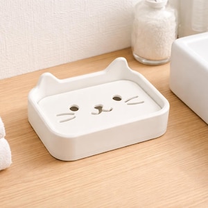 Op de afbeelding: Een wit, rechthoekig zeepbakje in de vorm van een kattenkop met twee kleine oortjes aan de bovenrand. Het oppervlak is voorzien van uitsparingen in de vorm van twee ronde ogen, een neusje en drie snorharen aan weerszijden voor afwatering.