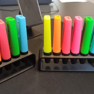 May include: Two black pen holders each holding a set of colorful highlighters. The highlighters are in shades of orange, pink, green, and blue. The highlighters are cylindrical with a rectangular tip. The holders are on a dark surface.