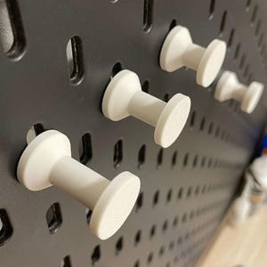 Peut inclure: Gros plan sur un panneau perforé noir avec plusieurs crochets de panneau perforé blancs insérés. Les crochets ont un corps cylindrique et une tête plate et circulaire. Le panneau perforé comporte des trous espacés pour les crochets. Quelques bouteilles sont visibles en arrière-plan.